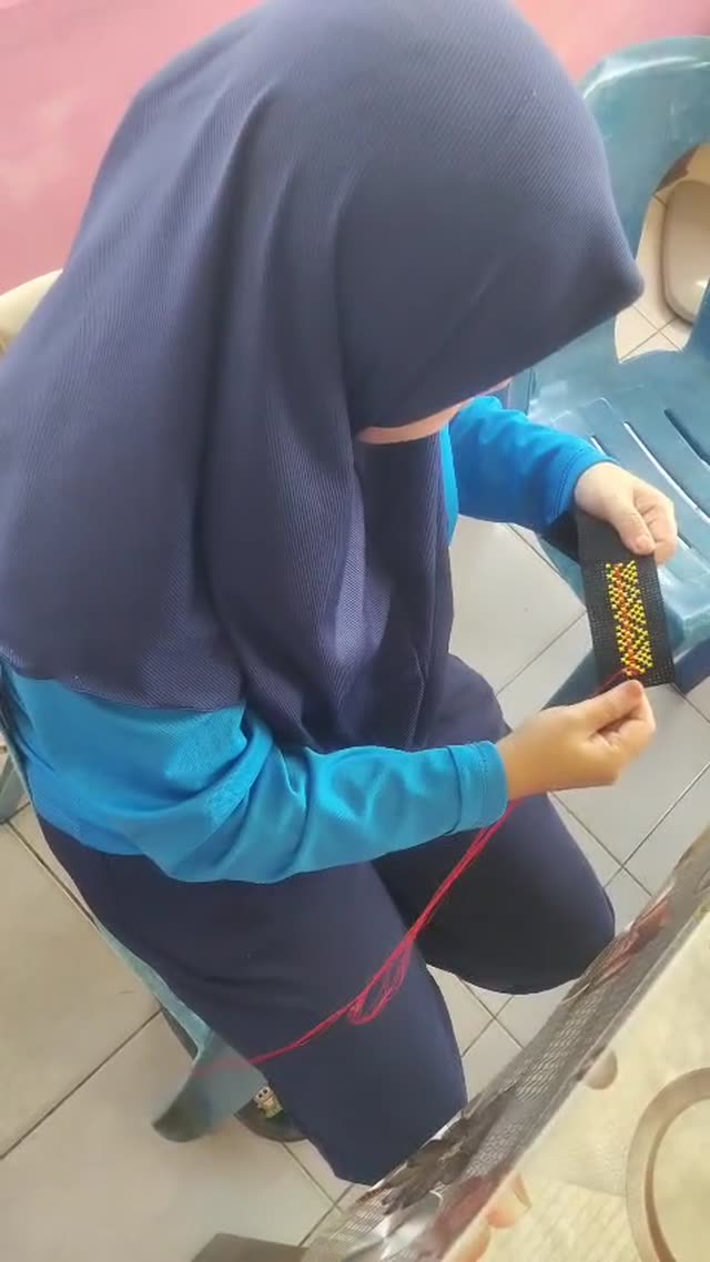 Close-up tangan menusang benang merah ke dalam anyaman hitam untuk membentuk corak kuning dan merah